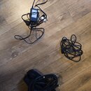 Na dřevěné podlaze leží tři černé kabelové komponenty. V horní části je síťový adaptér s připojeným kabelem a vidlicí. Vpravo je srolovaný černý kabe…