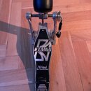 Fotografie znázorňuje basový pedál v černé a stříbrné barvě. Na černém základním plátu je vidět bílé logo TAMA a nápis The Legend in Innovation. Pedá…