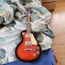 Elektrická kytara s trupem v barevnosti sunburst a bílým pickguardem. Na hlavě hmatníku je viditelné logo Epiphone. Nástroj má dva humbuckery s krémo…