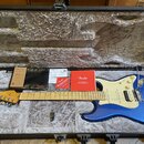 Modrá elektrická kytara s bílým pickguardem v otevřeném černém kufru s šedou výplní. Na hlavě kytary je logo Fender. V kufru jsou umístěny dokumenty…
