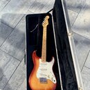 Elektrická kytara s tmelem v červeno-žlutém přechodu (sunburst) a bílým pickguardem v otevřeném hliníkovém kufru s černým polstrováním. Na hlavě hmat…