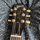 Fotografie zachycuje hlavu černé elektrické kytary s bílými ladicími kolíky. Na hlavě je bílý nápis Gibson a pod ním kurzivní nápis Les Paul s malým…