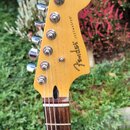 Fotografie zachicuje hlavu a hриф elektrické kytary s přírodním dřevěným povrchem. Na hlavě je vidět nápis Fender JAZZMASTER®. Hриф má tmavý hmatník…
