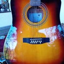 Akustická kytara s vyřezem v těle a povrchovým provedením sunburst (přechod od žluté k tmavě hnědé). V otvoru kytary je vidět modrý papírový štítek s…