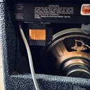 Záběr na zadní část kytarového komba s černým texturovaným povrchem. Je vidět reproduktor a štítek s nápisy „35 WATTS“, „16.7V RMS“, „8 OHMS“ a „220/…