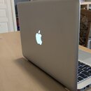 Stříbrný notebook s hliníkovým povrchem, který je otevřený a položený na světlém dřevěném stole. Na vnější straně víka je umístěno bílé svítící logo…
