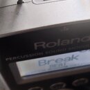 Stříbrný Percussion Sound Module značky Roland. Na horní straně je USB port a kovová rukojeť. Displej zobrazuje nápisy Break a BEAT.