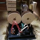 Fotografie znázorňuje vnitřní součásti elektronického zařízení s dvěma velkými bílými kruhovými cívkami. Na bílých obdélníkových součástkách je trojn…