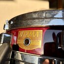 Detail červeného snare drumu s kovovými obručmi. Na korpusu je zlatá štítková nálepka s nápisy KITAMA, SNARE DRUM, SERIAL No. 049024 a MADE IN JAPAN.…