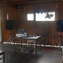 Fotografie zachycuje DJ set umístěný v dřevěné místnosti. Na stole se nachází notebook, bílý kufřík a DJ mixážní pult. Po stranách jsou dva černé rep…