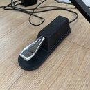 Černý sustain pedál s kovovým stříbrným šlapadlem. Na horní části těla je vyražen nápis YAMAHA. Pedál je umístěn na dřevěné podlaze vedle napájecího…