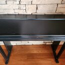 Černý dřevěný stolek pro klavír nebo digitální piano s výraznou strukturou žilování. Má obdélníkový tvar s mírně vyčnívající přední deskou a dvěma ši…