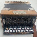 Černý harmonika s hnědými dřevěnými elementy a koženým popruhem. Na horní části je kovový štitek s nečitelným nápisem. Přední strana obsahuje ozdobno…
