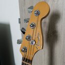 Fotografie zachycuje hlavu basové kytary z přírodního dřeva s čtyřmi chromovanými ladičkami. Na povrchu je bílý nápis Fender a menší text JAZZ BASS.…