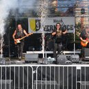 Fotografie zachycuje kapelu na pódiu s nástroji. Vlevo hraje kytarista s bílou elektrickou kytarou, vpravo kytarista s červeně stínovanou elektrickou…
