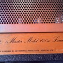 Detailní záběr na spodní část předního panelu kytarového komba s kovovou mřížkou. Na zlatavém kovovém pásu je nápis „Mk 2. Master Model 100w Lead.“ a…