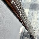 Detailní pohled na hmatník a krk basové kytary s tmavým dřevěným povrchem. Jsou vidět čtyři kovové struny a světlejší pražníky. Na hraně hmatníku jso…