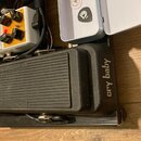 Černý wah-pedál s nápisem cry baby. Vedle něj je otevřená bílá plastová krabička s logem Dunlop a nápisem USA. V pozadí jsou vidět další efektové ped…