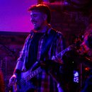 Fotografie zachycuje hudebníka s elektrickou basovou kytarou v tmavé barvě s modrým odleskem. Nástroj má čtyři struny a viditelný černý úhlový potenc…