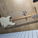 Elektrická basová kytara v krémové barvě položená na kartonovém obalu. Na zadní straně krku je vidět logo Squier by Fender. Tělo nástroje má v dolní…
