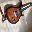 Elektrická kytara s tmelejším povrchem v barvě sunburst a viditnou strukturou dřeva. Nástroj má dva humbuckery, tři oranžové oválné knofíky a jeden p…