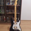 Černá elektrická kytara s bílým pickguardem a javorovým hnedým hmatníkem. Na hlavě je viditelné logo Fender. Kytara má tři snímače, pětistupňový přep…