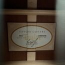Fotografie zachycuje vnitřní štítek akustické kytary přilepený k dřevěnému korpusu. Na štítku je uveden nápis TAYLOR GUITARS, model 326ce a SERIAL NO…