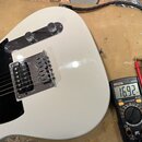 Bílá elektrická kytara s černým pickguardem a chromovaným hardwarem. Na těle je vidět kobylka s hmyvkami a dvě chromované knoflíky. Vedle kytary leží…