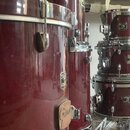 Fotografie zobrazuje několik červených bubnů s chromovaným kováním. Na korusech jsou viditelné loga GRETSCH a nápisy "Custom Magic". Povrch nástrojů…