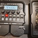 Černý kytarový procesor s displejem, na kterém je nápis 24 RocknRoll. Na horní části jsou nápisy G1X FOUR, ZOOM a Multi-Effects Processor. Zařízení m…