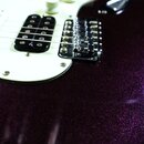 Detail elektrické kytary s tmavě fialovým metalizovaným povrchem s třpytivým efektem. Nástroj má bílý pickguard, tři snímače (dva jednoduché a jeden…