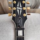 Černá hlava elektrické kytary s bílým lemováním a zlatými ladičkami. V horní části je nápis Epiphone, uprostřed je vložka ve tvaru kosočtverce a níže…