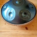 Kulatý steel tongue drum v tmavě modré barvě s několika vyhlubecinami na povrchu. Nástroj je umístěn v pleteném hnědém obalu z přírodního materiálu.…