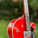 Červená polodublová elektrická kytara s bílým lemováním hran. Na těle jsou vidět dva stříbrné humbuckery a černý plecháč. Povrch laku je lesklý, bez…