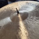 Záběr na povrch zlatavého činchu s viditelným broušením a kladivováním. V horní části je částečně viditelný nápis Istanbul Agop Handmade Cymbals. Upr…