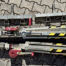 Na fotografii jsou rozložené kovové konstrukce ständerů v stříbrné barvě s červenými prvky a žlutými varovnými pruhy. Jsou vidět dvě základny s úchyt…