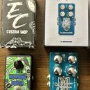 Fotografie zobrazuje dva kytarové efekty s jejich originálními krabicemi. Jeden pedál je zelený s nápisy HOT RODDED BOOSTER a EC CUSTOM SHOP, druhý j…