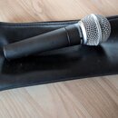Černý dynamický mikrofon s kovovou síťkovou hlavicí v barvě stříbra. Na těle mikrofonu je bílý nápis SHURE SM58. Mikrofon leží na černém koženěném ne…