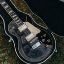 Černá elektrická kytara s tvarem single-cut v otevřeném černém kufru na trávě. Nástroj má dva humbuckery s krémovými kryty, krémový pickguard a čtyři…