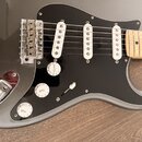 Šedá elektrická kytara s černým pickguardem a třemi bílými snímači. Na ovládacích knoflících jsou nápisy VOLUME a TONE s číslicemi 1 až 10. Na snímač…