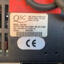 Detail štítku na zadní straně černého elektronického zařízení. Na bílém štítku je logo QSC a text: QSC AUDIO PRODUCTS COSTA MESA, CA USA MADE IN USA…
