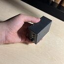 Ruka drží malý, černý obdélníkový box z kovu s matným povrchem. Na jedné straně jsou vidět dva malé šrouby a jeden konektor typu jack. Na povrchu nej…