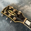 Hlava elektrické kytary v černé barvě se zlatým lemováním. Na hlavě je bílé logo Gibson a nápis SG CUSTOM. Jsou vidět kovové ladičky a zlatý ornament…