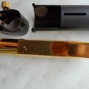 Fotografie zobrazuje tři části příslušenství k dechovému nástroji. V horní části je černý plastový munčnípíst s nápisem 19 a černá ligature z karbono…