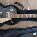 Černá elektrická kytara s tvarem single-cut v otevřeném černém kufru. Na hlavě je bílý nápis Epiphone a stylizovaný nápis Les Paul. Kytara má dva hum…