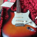 Elektrická kytara s tělem v provedení sunburst a bílým pickguardem leží v červeném plišovém kufru. Na hlavě hředu je viditelné logo Fender. Kytara má…