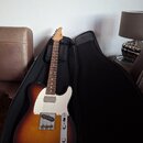 Elektrická kytara s tělem v barvě sunburst a bílým pickguardem, umístěná v černém obalu. Na hlavě hmatníku je viditelné logo "Tele". Kytara má jeden…