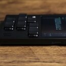 Černý elektronický přístroj s displejem a několika ovládacími tlačítky, položený na dřevěném povrchu. Na displeji je vidět tabulka s textem a hodnota…