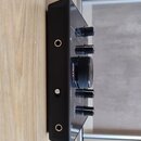 Černý elektronický přístroj s pětí knoflíky, z nichž prostřední je výrazně větší a rýhovaný. Na středovém knoflíku je červeným písmem nápis "ALL". Po…