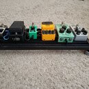 Na černém podložkovém pedalboardu s kolečky je umístěno pět efektů. Zleva jsou vidět pedály s nápisy: neoznačený kovový pedal, Cry baby DUNCAN, zelen…
